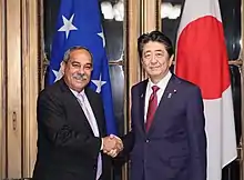 Deux hommes souriant, placés devant les drapeaux de leurs pays respectifs, se serrent la main.