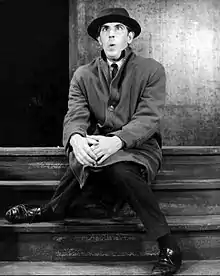 Photo en noir et blanc d'un homme en costume, manteau et chapeau, assis sur une marche d'escalier et qui fait une grimace amusante.