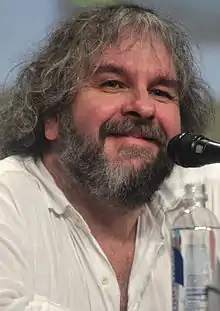 Photographie en couleur d'un homme souriant devant un microphone, aux cheveux et à la barbe poivre et sel, portant un chemisier blanc.