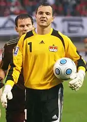 Un gardien de but de football avec un maillot jaune tient un ballon. Juste derrière lui, il y a un autre homme.