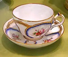 Une tasse en porcelaine avec sa soucoupe, blanches à liserés dorés et décorations bleues.