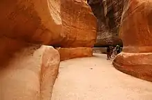 Couloir sinueux creusé dans la roche avec deux rigoles latérales également creusées dans la roche. Quelques personnes marchent en arrière plan au fond du couloir.