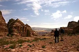 Vue de loin d'un monument sculpté dans la roche. Une esplanade de roche et de sable se trouve en avant plan. Tout au fond, il y a un paysage désertique.
