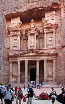 Un monument sculpté dans la roche. Le soleil illumine la moitié supérieure du monument et des gens en sortent. Il y a un chameau assis devant l'édifice.