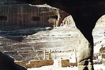 Gradins d'un théâtre romain, avec arrière plan de rochers. Des ouvertures approximativement rectangulaires sont pratiquées dans la roche. En avant plan est partiellement occupé par l'entrée vue de l'intérieur d'une cavité à partir de laquelle la photo a été prise.