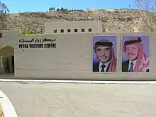 Photographie d'un mur moderne pourvu d'une enseigne en anglais et en arabe. Sur la gauche du mur, il y a une entrée vitrée et, sur la droite, deux affiches représentent chacune le visage d'un homme différent. Ils sont tous deux coiffés d'un keffieh. Une rue passe devant le mur.
