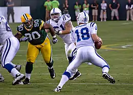 De dos, un joueur numéro 18 en maillot blanc, s'appuie sur son pied gauche pour fuir un défenseur en vert et jaune qui court vers lui, sur un terrain de football américain.