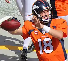 Un joueur de football américain avec un casque bleu foncé et un maillot orange, ballon en main et l'autre main en l'air pour préparer son geste de lancer.