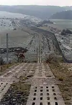 Une route de patrouille composée de deux bandes parallèles de blocs de béton perforés descend dans une vallée le long d'une pente raide. À droite, une clôture continue longe la route. La route et la clôture se prolongent dans un paysage légèrement enneigé jusqu'à une colline boisée.