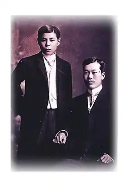 Deux hommes asiatiques vêtus à l'occidentale, l'un debout, âgé d'une vingtaine d'années, l'autre un peu plus vieux, assis.