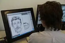 Photo couleur d'une personne, vue de dos, assise devant un écran d'ordinateur dans lequel un logiciel de dessin est ouvert et affiche le portrait noir et blanc d'un homme barbu.