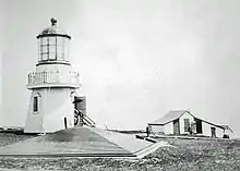 photo noir et blanc avec le phare à gauche des petits bâtiments à droite et la toiture à quatre pans, posée sur le sol, de la citerne à eau