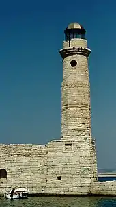 Un haut phare en pierre claire par temps ensoleillé en bas duquel mouille une barque blanche.