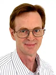 Photographie d'un homme roux, avec des lunettes, et portant une chemise blanche avec de fines rayures noires. Il regarde l'objectif.