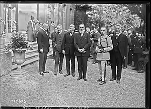photographie en noir et blanc de six hommes devant une foule au pied d'un escalier située à gauche.