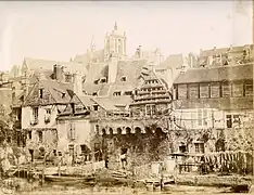 Photographie ancienne d'une ville très dense avec une muraille crénelée au premier plan, des maisons très dégradées et un clocher à l'arrière-pan