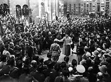 Photographie en noir et blanc d'un rassemblement de nombreux hommes, devant une église ; au centre deux hommes, l'un en uniforme est au garde-à-vous, l'autre lui tend des papiers. La foule est constituée de personnes soit en uniforme de soldat, casque de combat, soit en uniforme noir et toque noire.