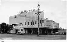 Photographie noir et blanc d'un grand bâtiment art-déco. Des lettres forment le nom du cinéma, Piccadilly, sur le toit du bâtiment.