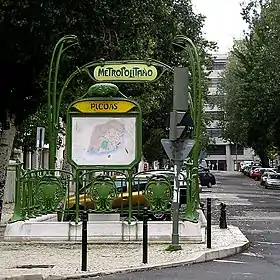 Photo couleur dans une rue arborée d'une balustrade en fer forgé à écussons et portique vu de l'arrière, les panneaux montrant les mots "Metropolitano" et "Picoas"