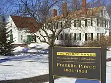 Photographie d'une maison blanche à deux étages avec de hautes cheminées en brique. Le sol est recouvert de neige et un panneau indique qu'il s'agit de la résidence du 14e président des États-Unis.