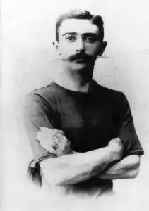 Photographie en noir & blanc de Pierre de Coubertin. Il croise les bras et porte une longue moustache.