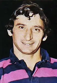 Photographie en couleurs. Portrait d'un homme jeune aux cheveux bouclés portant un polo rayé rose et violet.