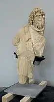 Vue d'une statue en pierre d'un personnage barbu, sans bras ni jambes, coiffé d'un bonnet phrygien.