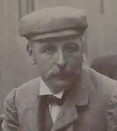 Photo en portrait d'un homme arborant une moustache en brosse et un couvre-chef.