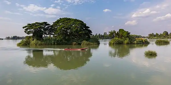 Pirogue filant sur le Mékong devant une petite île avec des arbres verts se reflétant dans l'eau, dans le Si Phan Don. Avril 2018.