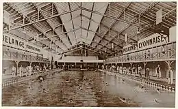 Vue du grand bassin de la piscine avec des nageurs sous un toit du type halle. Au fond du bassin sur le petit côté se trouvent des gradins.