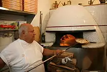 Photo d'un pizzaiolo en train de retirer une pizza d'un four à bois à l'aide d'un long manche. On voit le bois brûler à l'intérieur du four à travers son ouverture. L'extérieur du four est en céramique blanche, des buches de bois sont entassé au pied du four.