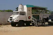 Photo d'un camion blanc aménagé pour produire et vendre des pizzas à l'arrière. Trois marches posées au sol permettent d'accèder au comptoir sous un auvent. Sur le flanc du camion, on peut lire « Pizza à emporter » avec un numéro de téléphone, et « 1 gratuite Pour 9 achetées ».