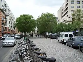 Photo en couleurs d'une place rectangulaire à large terre-plein central avec bicyclettes au premier plan, arbres au fond, et immeubles de part et d'autre