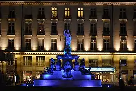 La fontaine éclairée en bleu, se détachant devant les façades en beige et ocre.