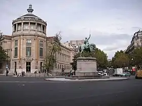 Photographie en couleur d'une place ornée d'une statue de cavalier, à gauche un immeuble en rotonde, à droite une avenue