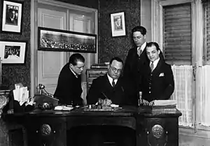 Photographie en noir et blanc de quatre hommes derrière un bureau. L'un d'eux écrit sur un document.