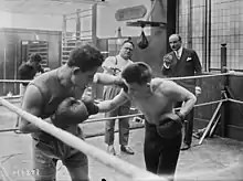 Photographie en noir et blanc de deux boxeurs s'entraînant dans un ring, à l'extérieur de celui-ci deux hommes les observent et font des gestes.