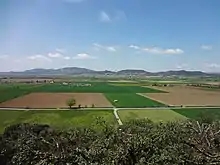 D'un point de vue en hauteur, on voit la plaine découpée en champs carrés de différentes couleurs. Au fond, petites montagnes dans le lointain, et ciel bleu moucheté de nuages.