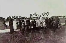 Photo en noir et blanc d'un avion sur le dos, entouré de plusieurs personnes.