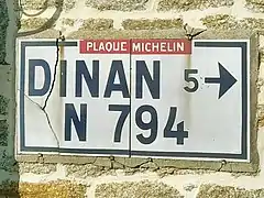 Plaque Michelin scellée sur le mur d’une maison et indiquant la ville de Dinan à 5 kilomètres sur la route de Corseul (D794).