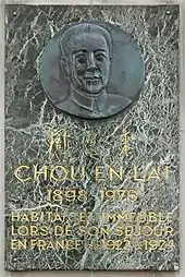 Photographie d'une plaque de marbre portant en haut un buste sculpté sur un médaillon métallique, au centre trois sinogrammes et en bas le texte suivant : « Chou En Laï 1898-1976 habitat cet immeuble lors de son séjour en France de 1922 à 1924 ».
