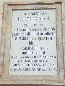 Plaque de marbre accrochée à un mur de pierre. Des phrases en latin y sont gravées.