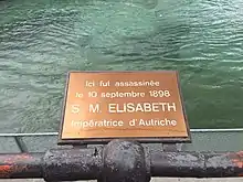 Photographie en couleurs d'une plaque de cuivre rappelant le lieu de l'assassinat de l'impératrice Élisabeth en face d'une étendue d'eau.