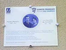 Plaque blanche portant en bleu la photo du couple Grandou jeunes, les logos de Mayrac et du Comité français Yad Vashem et une exhortation des passants à se souvenir.