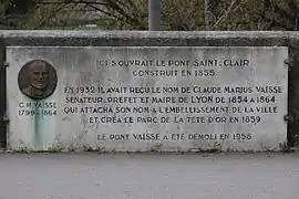 Photographie d'une plaque fixée sur un parapet, avec un texte et un médaillon représentant un visage.