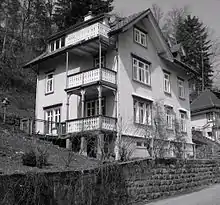 Photo noir et blanc d'une grande maison de trois étages style 1900, avec des balcons et des grandes fenêtres, forêt en arrière-plan.