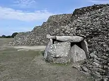 Énormes dalles debout côte à côte formant un polygone, et supportant une grande table. En fond, l'arrière du cairn secondaire, vu en coupe dans la zone dévastée, forme un escalier à trois marches.