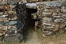 Vue de face de l'avant-couloir et de l'entrée, qui a une grille. Au fond du couloir, on voit le jour et un orthostate de la chambre éventrée.