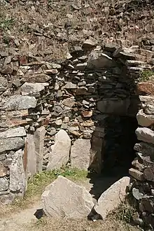 Vue de trois quarts d'une chambre en encorbellement éventrée. Des dalles sont plantées debout, en cercle, certaines incorporées dans la muraille. On aperçoit au fond à droite le débouché du couloir.