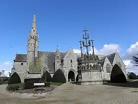 Vue du sud. Le porche ouest est à deux niveaux. Le clocher-mur est de profil. Vient ensuite le porche ouest, à deux niveaux et flanqué d'une tourelle. Deux petites chapelles sont masquées par le calvaire. Puis on voit une petite chapelle et une grande.